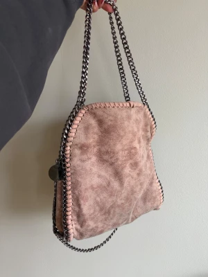 Rosa axelväska Falabela - Säljer en snygg axelväska i rosa mockaliknande material med grova silverfärgade kedjeremmar. Väskan har dekorativa sömmar längs kanterna. En av sömmarna har dragits ur ena hålet men går nog att peta in igen med tålamod. Super bra skick