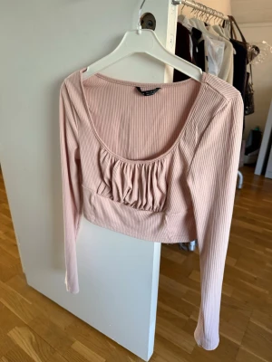 Ljusrosa croppad topp med rynk - Trendig ljusrosa croppad topp med långa ärmar och ribbad struktur. Toppen har en djup rundad urringning och snygga rynkdetaljer framtill. Perfekt att styla med höga jeans eller kjol för en cool look.