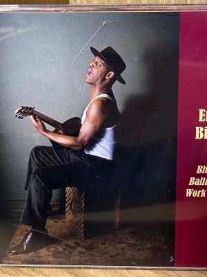 Eric Bibb Blues Ballads & Work Songs CD - Säljer en CD med Eric Bibb, 'Blues, Ballads & Work Songs'. Albumet innehåller 14 spår med blues och ballader. Omslaget har en bild på Eric Bibb med gitarr, klädd i vit linne och svart hatt. Perfekt för dig som gillar akustisk blues och genuina låtar.