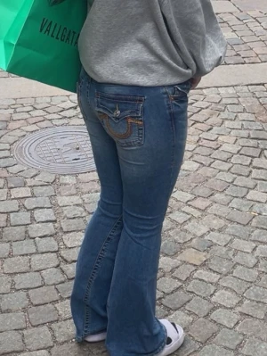 Blå bootcut jeans från True Religion - Säljer ett par blå bootcut jeans från True Religion med ikoniska broderade fickor bak och snygga kontrastsömmar. Jeansen har låg midja och klassisk femficksdesign. Perfekta för dig som gillar en avslappnad men trendig stil. Dragkedjan är lite lurig 