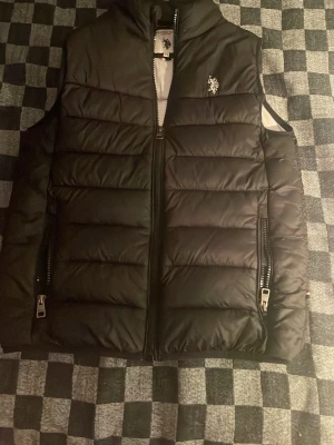 Svart dunväst från U.S. Polo Assn. - Snygg svart dunväst från U.S. Polo Assn. med quiltad design och dragkedja framtill. Västen har två fickor med dragkedja och en broderad logga på bröstet. Perfekt för lager-på-lager och streetstyle. Perfekt till vårens milda dagar