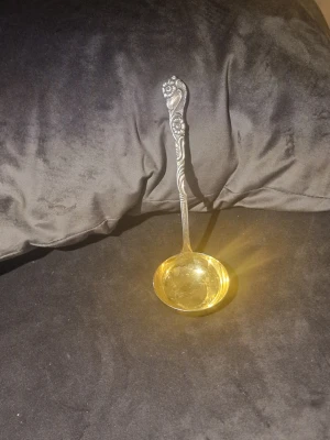 Dekorativ nysilversked med blommönster - Säljer en dekor crossad soppslev i silverfärg med detaljerat blommönster på handtaget. Skeden har en rund, djup skål och ett elegant, snirkligt handtag. Perfekt för servering av soppa eller sås och ger en klassisk touch till dukningen.