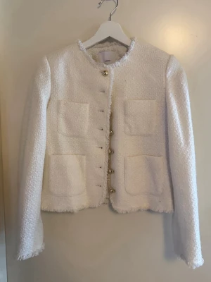 Vit bouclé kavaj från Mango - Stilren vit bouclé kavaj från Mango med rund halsringning och dekorativa guldknappar framtill. Kavajen har fyra stora fickor på framsidan och råa kanter för en trendig look. Perfekt att styla till jeans eller kjol för en chic outfit.