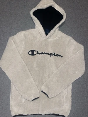 Beige fluffig hoodie från Champion - Mysig beige hoodie från Champion i fluffigt teddy-material. Stor broderad Champion-logga i svart på bröstet och svart innerfoder i huvan. Perfekt för dig som vill ha en skön och stilren hoodie med streetwear-vibe.