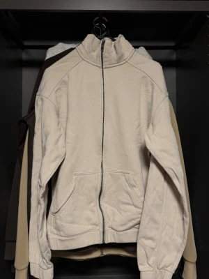 Beige zip hoodie med fickor - En stilren beige hoodie med dragkedja framtill och två stora fickor. Hoodien har en hög krage och ribbade muddar vid ärmar och nederkant. Perfekt för en avslappnad look och enkel att matcha med andra plagg.