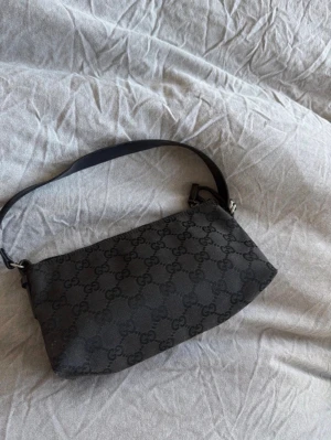Svart axelremsväska från Gucci - Snygg svart handväska från Gucci med klassiskt GG-mönster över hela väskan. Väskan har en enkel, rektangulär form och en bred axelrem i svart läder. Materialet är canvas med läderdetaljer och dragkedja upptill. Perfekt för dig som gillar stilrena designer. Pris går att diskutera 
