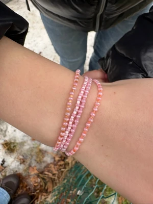 Rosa pärlarmband  - Säljer ett set med tre elastiska armband gjorda av små glaspärlor i olika nyanser av rosa. Armbanden är tunna och kan bäras tillsammans eller separat för en söt och färgglad look. Perfekt accessoar för att addera lite färg till din outfit.