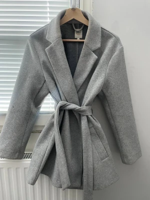 Grå kappa med knytskärp från ONLY - Snygg grå kappa från ONLY med klassisk krage och knytskärp i midjan. Kappan har raka ärmar och två sidofickor. Materialet är mjukt och känns som ullblandning, perfekt för kyliga dagar. Stilren och enkel design som passar till många outfits.