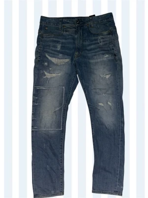 G-star Raw jeans - Säljer ett par blå jeans med slitningar och patchdetaljer på framsidan. Jeansen har klassisk femficksdesign och raka ben. Materialet är denim och färgen är en tvättad blå nyans med distressed look. Perfekt för dig som gillar en cool och avslappnad stil. Storlek 29/30.