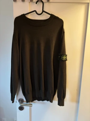 Mörkgrön Stone Island tröja - Säljer en mörkgrön tröja från Stone Island med den klassiska avtagbara logotypen på ärmen. Tröjan har rund halsringning, ribbade muddar och är tillverkad i mjukt stickat material. Perfekt för dig som gillar streetwear och vill ha en clean look.