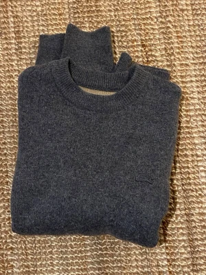 Grå Lacoste Stickad Tröja (Nyskick) - Säljer en riktigt snygg och stilren stickad tröja från Lacoste i en mörkgrå melerad färg. Tröjan har den klassiska krokodilloggan broderad på bröstet i en diskret matchande färg (ton-i-ton), vilket ger ett exklusivt intryck. Tröjan är oanvänd utan prislapp, 10/10 skick. Storlek M.