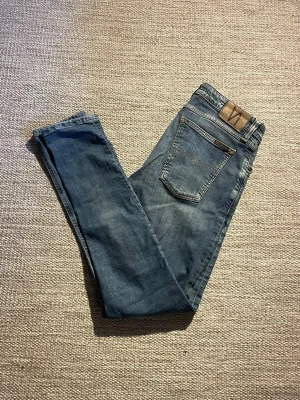 Blå jeans från Nudie Jeans - Säljer ett par klassiska blå skinny jeans från Nudie Jeans. Jeansen har fem fickor, normal midja och är tillverkade i mjuk denim med lätt slitningar. Snygg läderpatch med logga bak i midjan och smal passform som sitter tajt hela vägen ner.