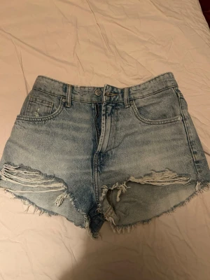 Ljusblå denimshorts med slitningar - Säljer ett par ljusblå denimshorts med råa kanter och slitna detaljer framtill. Klassisk femficksmodell med bälteshällor och knappgylf. Perfekta för sommaren och ger en avslappnad vibe.