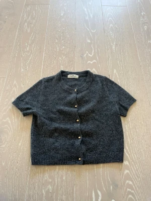 Mörkgrå kofta från H&M - Kortärmad mörkgrå kofta från H&M med rund halsringning och knappar framtill. Koftan är stickad i ett mjukt material och har ribbade kanter vid ärmslut och nederkant. Perfekt att slänga över en topp för en chill look.