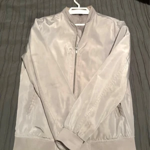 Ljusgrå bomberjacka från Zara Man - Stilren ljusgrå bomberjacka från Zara Man i blankt, lätt material. Jackan har dragkedja framtill, två sidofickor och ribbade muddar vid ärmslut och nederkant. Perfekt för dig som gillar minimalistisk och modern stil.