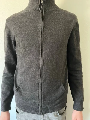 Mörkgrå ziptröja med hög krage - Mörkgrå ziptröja med hög krage och hel dragkedja framtill. Tröjan har två fickor och ribbade muddar vid ärmslut och nederkant. Perfekt för lager-på-lager och enkel att matcha med olika outfits.