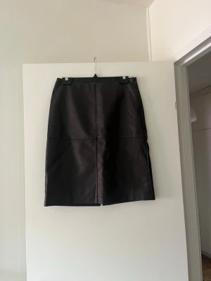 Svart långkjol i skinnimitation - Snygg svart långkjol i skinnimitation med raka linjer och minimalistisk design. Kjolen har markerade sömmar framtill som ger en cool touch. Perfekt att styla med chunky boots eller en stickad tröja.