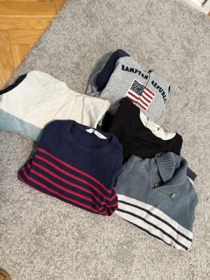 5-pack tröjor & koftor, olika färger - Säljer ett paket med fem tröjor och koftor i olika färger och stilar. Inkluderar en marinblå tröja med röda ränder, en grå kofta med vita ränder och knappar, en grå ziptröja med tryck, samt två t-shirts i vitt och svart. Perfekt för dig som vill variera din stil.