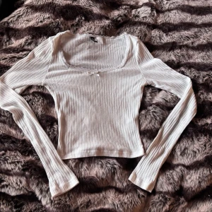 Vit ribbad topp med spetsdetalj från hm - Söt vit långärmad topp i ribbat material med fyrkantig urringning. Dekorerad med spetskant och liten rosett framtill. Stretchig och figurnära passform, perfekt till jeans eller kjol.