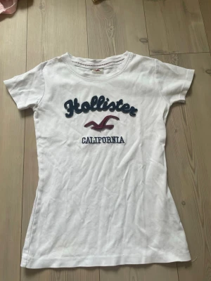 Vit Hollister t-shirt med tryck - Säljer en vit t-shirt från Hollister med blå och röd broderad logga och texten 'California' på bröstet. T-shirten har rund hals, korta ärmar och är i mjuk bomull. Passformen är normal och den är perfekt för en avslappnad stil.