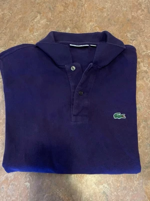 Mörkblå piké från Lacoste - Säljer en klassisk mörkblå pikétröja från Lacoste med den ikoniska gröna krokodilloggan på bröstet. Tröjan har korta ärmar, krage och knappar framtill. Tillverkad i mjuk bomull som känns skön mot huden.