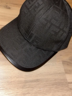 Svart keps från Fendi med logomönster - Snygg svart keps från Fendi med diskret ton-i-ton logomönster över hela kepsen. Justerbar rem bak med metallspänne och klassisk böjd skärm. Tillverkad i ett slitstarkt tyg som känns exklusivt och trendigt.
