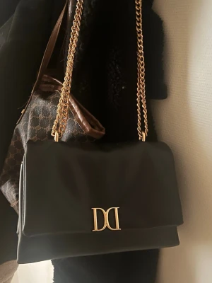 Svart handväska från Don Donna - Stilren svart handväska från Don Donna med guldfärgad kedjerem och DD-logga framtill. Väskan har en klassisk rektangulär form och är tillverkad i slätt konstläder. Perfekt accessoar för att lyfta din outfit.