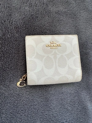 Coach beige plånbok med monogram - Snygg beige plånbok från Coach med klassiskt monogrammönster och guldfärgade detaljer. Plånboken har dragkedjefack, flera kortfack och en knappstängning i brunt läder. Perfekt storlek för fickan eller väskan.