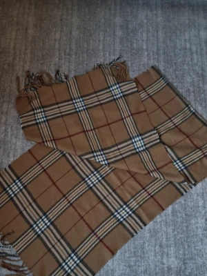 Burberry liknande halsduk  - Klassisk beige halsduk med rutigt mönster i svart, vitt och rött. Halsduken har fransar i ändarna och är tillverkad i ett mjukt tyg som känns skönt mot huden. Perfekt accessoar för att lyfta din outfit med en tidlös look.