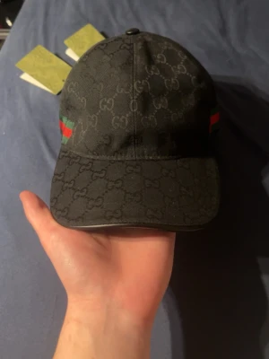 Svart Gucci keps med GG-mönster - Snygg svart keps från Gucci med klassiskt GG-monogram över hela kepsen. På sidan finns det ikoniska gröna och röda bandet. Kepsen har böjd skärm och justerbar rem bak. Perfekt för dig som vill ha en exklusiv och trendig accessoar.