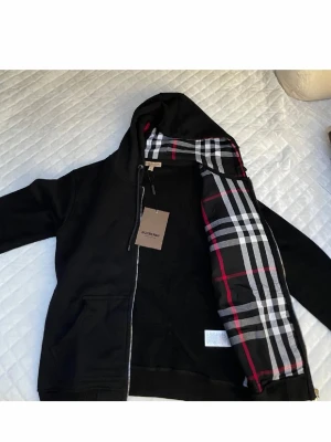 Svart hoodie från Burberry - Snygg svart hoodie från Burberry med dragkedja och klassiskt rutigt foder i huvan. Hoodien har fickor framtill och justerbar dragsko i huvan. Perfekt för dig som vill ha en stilren och exklusiv look med en twist av det ikoniska Burberry-mönstret.
