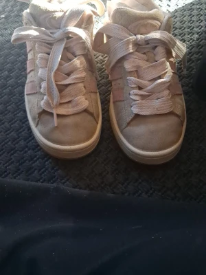 Beige Adidas sneakers med rosa ränder - Säljer ett par snygga Adidas sneakers i beige mocka med klassiska rosa ränder på sidorna. Skorna har vita breda skosnören och låg profil. Perfekta för dig som gillar en stilren och sportig look.