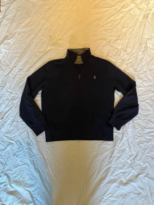 Mörkblå half-zip tröja Polo Ralph Lauren - Mörkblå tröja från Polo Ralph Lauren med halv dragkedja och ståkrage. Klassisk broderad logga på bröstet i vitt. Insidan av kragen är ljusgrå. Perfekt för en avslappnad men stilren look.