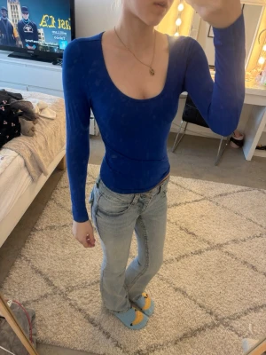 Blå långärmad topp med djup ringning - Säljer en tight, långärmad topp i klarblå färg med djup rund ringning. Toppen har en slim passform och är tillverkad i ett stretchigt material som sitter snyggt på kroppen. Perfekt att styla med jeans eller kjol för en trendig look.