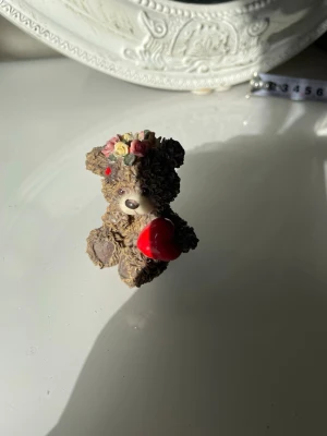 Vintage nallefigur med rött hjärta - Dekorativ liten nallebjörn i brunt konstmaterial som håller ett rött hjärta. Nallen har detaljer som blommor i olika färger på huvudet och en söt nos. Perfekt som prydnad eller present till någon speciell.gjort av sten eller nåt annat vet inte exakt 