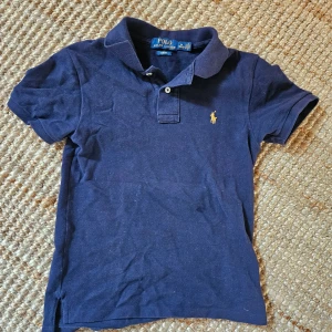 Marinblå pikétröja från Ralph Lauren - Klassisk marinblå pikétröja från Ralph Lauren med korta ärmar och krage. Stl M (10-12). Tröjan har den ikoniska broderade loggan i gult på bröstet och är i slim fit-modell. Perfekt till jeans eller shorts för en stilren look.