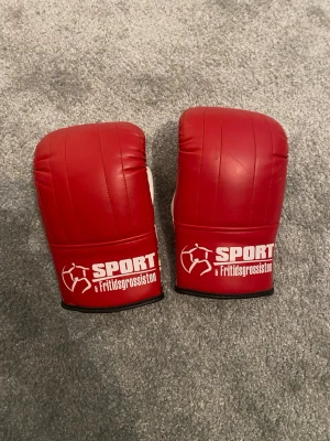 Röda boxningshandskar Sport & Fritidsgrossisten - Säljer ett par röda boxningshandskar från Sport & Fritidsgrossisten. Handskarna har vit insida och svart kant, med vadderad ovansida och kardborreknäppning vid handleden. Perfekta för träning eller sparring inom kampsport.