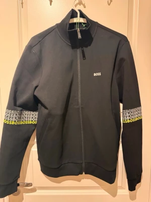 Svart zip hoodie från BOSS - Svart hoodie från BOSS med hel dragkedja och ribbade muddar. Diskret BOSS-logga på bröstet och upprepade BOSS-tryck i vitt och gult på båda ärmarna. Tillverkad i mjukt bomullsmaterial och har en modern, sportig look. Perfekt för dig som gillar streetwear. Helt ny med prislapp och oanvänd. 