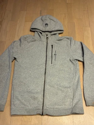 Sail Racing Zip Hoodie - Snygg ljusgrå hoodie från Sail Racing med dragkedja framtill och en bröstficka med svart logga. Hoodien har en broderad logga på huvan och är gjord i mjukt, fleecefodrat material. Perfekt för chill dagar och en clean-stil.