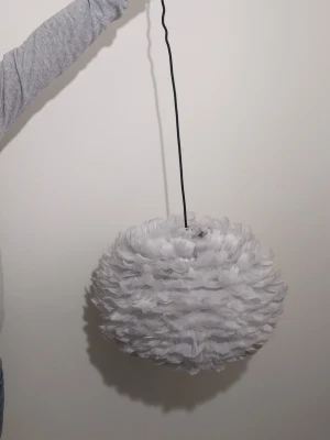 Vit fjäderlampa från Mio - Säljer en rund taklampa från Mio med vit lampskärm täckt av mjuka fjädrar. Lampan har svart sladd och ger ett mysigt och modernt intryck i rummet. Passar perfekt som statement piece i sovrum eller vardagsrum.