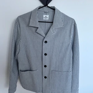 Randig pyjamasskjorta John Henric - Klassisk pyjamasskjorta från John Henric med smala vertikala ränder i vitt och mörkblått. Skjortan har en avslappnad krage, knäppning framtill och två fickor nedtill. Tillverkad i mjuk bomull för extra komfort.