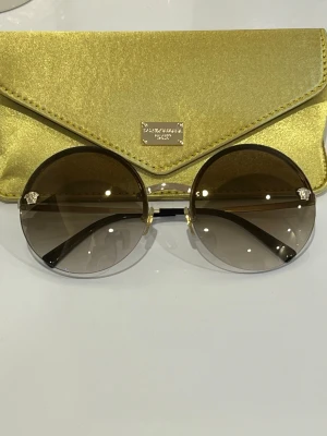 Versace runda solglasögon guld nyskick - Snygga runda solglasögon från Versace med tunna guldfärgade bågar och bruna tonade glas.                      Obs! Jag råkade blanda ihop glasögonfodralen på bilden. Det gröna fodralet tillhör Dolce & Gabbana-glasögon. Jag upptäckte först nu att det inte hör till just dessa glasögon. Jag har därför lagt till en bild på det korrekta, originalfodralet som är svart.  Vid köp kommer glasögonen att skickas med det originala svarta fodralet som ni kan se på bilden.