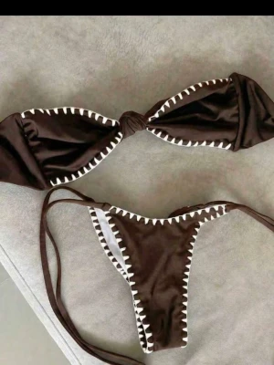 Brun bikini med vita detaljer - Snygg brun bikini med vita, sicksack-formade kanter. Överdelen är bandeau-modell med knut framtill och underdelen har smala knytband i sidorna. Perfekt för stranden eller poolen och ger en trendig look.