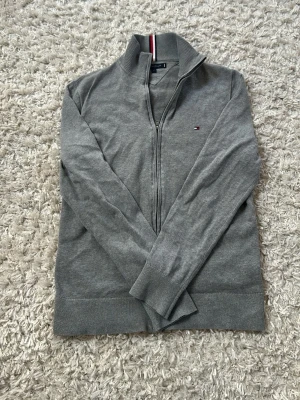 Grå ziptröja från Tommy Hilfiger - Snygg grå ziptröja från Tommy Hilfiger med liten logga på bröstet och ribbade muddar. Tröjan har hög krage med dragkedja och en diskret röd, vit och blå rand på insidan av kragen. Perfekt till lager på lager och enkel att matcha.