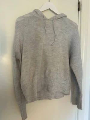 Ljusgrå stickad hoodie - Mysig ljusgrå stickad hoodie med huva och dragsko. Tröjan har ribbade muddar vid ärmslut och nederkant samt en avslappnad passform. Perfekt för dig som gillar en enkel och stilren look. Materialet känns mjukt och behagligt mot huden.