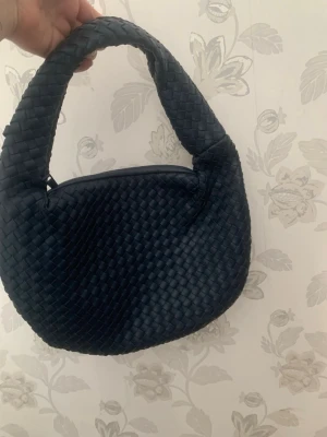 Mörkblå Väsk  axelväska i läderstil - Trendig mörkblå handväska med rundad form och bred axelrem. Väskan har ett snyggt flätat mönster över hela ytan och stängs med dragkedja. Perfekt accessoar för dig som gillar stilrena detaljer och modern design.