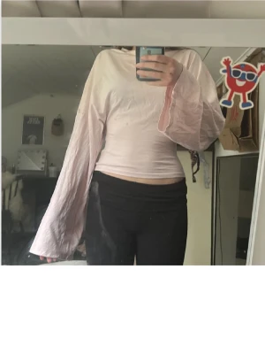 Ljusrosa topp med vida ärmar - Trendig ljusrosa topp med rund hals och extra vida, långa ärmar. Toppen har en figurnära passform i midjan och är tillverkad i ett mjukt bomullsliknande material. Perfekt för dig som vill sticka ut med en unik siluett.