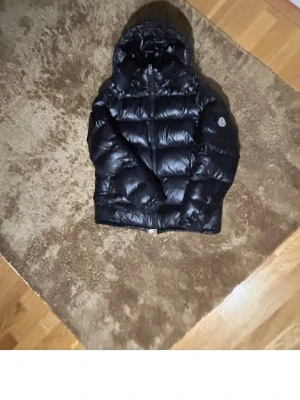 Pufferjacka med huva - Snygg svart pufferjacka  med huva och quiltad design. Jackan har dragkedja framtill och logga på ärmen. Perfekt för kalla vinterdagar och riktigt varm tack vare dunfyllningen.