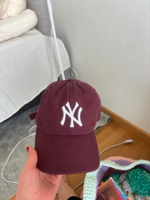 Vinröd New York keps  - Snygg vinröd keps från 47 Brand med klassisk New York Yankees-logga broderad i vitt framtill. Justerbar rem baktill för perfekt passform. Tillverkad i bomullstyg som känns både bekvämt och stilrent.