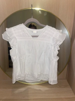 Vit blus med volangärmar från ginatricot  - Söt vit blus med v-ringning och volangdetaljer längs ärmarna. Blusen har ett luftigt tyg och fina spetsinslag upptill. Perfekt för en fräsch och somrig look. Materialet känns lätt och bekvämt.den har en lite sminkfläck på baksidan(se bild) men annars är den i toppskick!skriv för fler bilder,pris kan diskuteras!💗